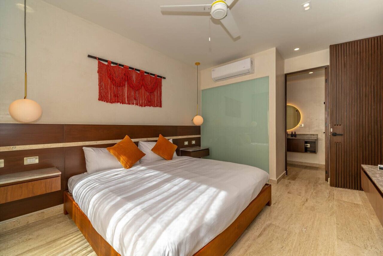 A modern hotel room with a large bed, wooden headboard, two bedside tables, a red decorative wall hanging, an air conditioning unit, a ceiling fan, and a view of the bathroom.