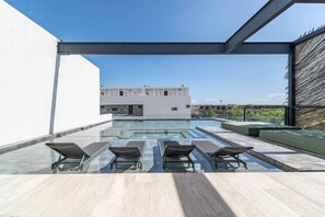 A rooftop swimming pool with glass railings and lounge chairs, a modern building in the background, and a clear blue sky.