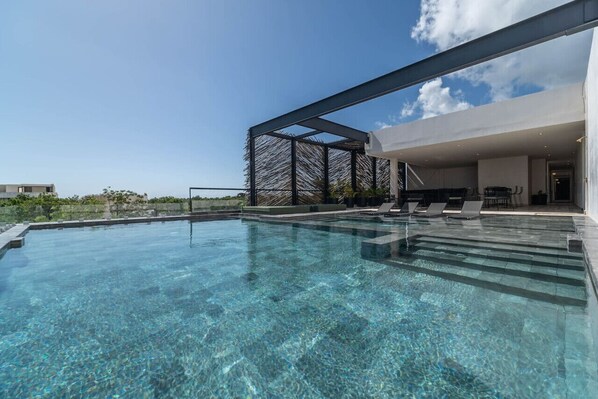 A clear swimming pool with steps leading into it, surrounded by a modern outdoor area with lounge chairs and a covered seating area. - Lux 1BR w/ Rooftop Pool, Close to Beach (Tulum)