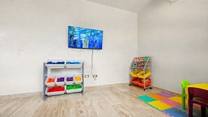 A room with a television mounted on the wall, a bookshelf with colorful storage bins, and a play area with a mat and a chair. - Lux 1BR w/ Rooftop Pool, Close to Beach (Tulum)
