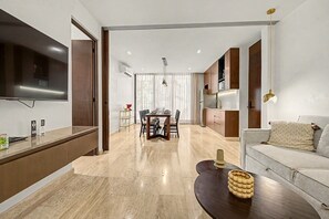 A modern living room with a flat-screen TV, a dining area with a table and chairs, and a kitchen with wooden cabinets and a built-in oven.