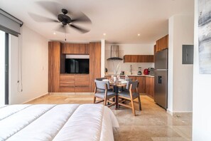 A modern hotel room with a bed, a dining area with chairs and a table, a kitchen with a refrigerator, and a ceiling fan. - King Suite w/ Balcony, Gym + Rooftop Pool (Tulum)