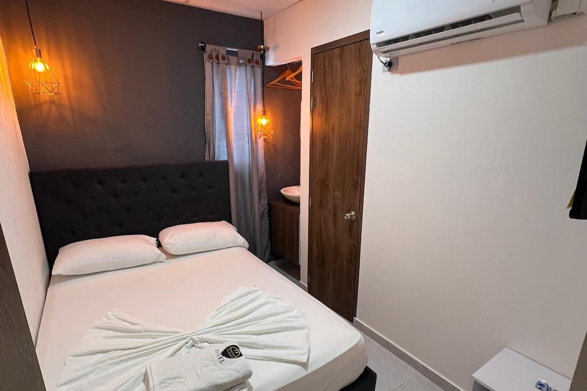A hotel room with a bed, a bedside table, a wardrobe, a bathroom, and an air conditioning unit.