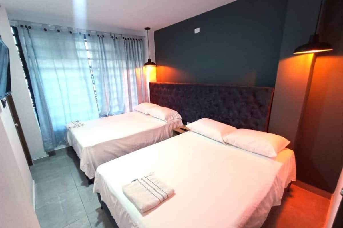 A hotel room with two beds, a dark headboard, a large window with curtains, and a wall-mounted lamp.
