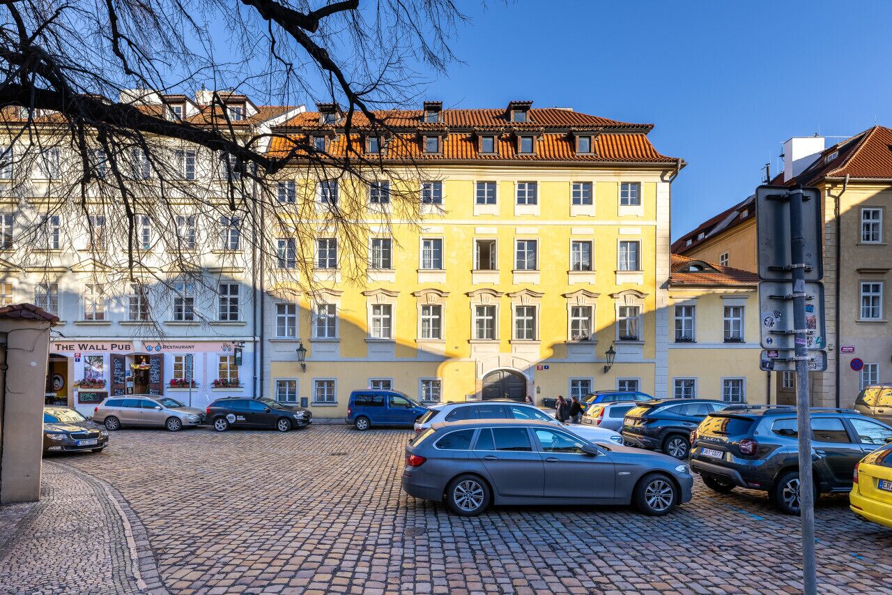 En brostensbelagt gade med parkerede biler og en bygning med gul facade og mange vinduer.