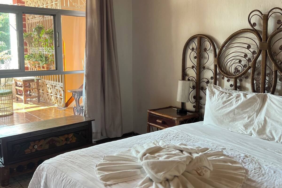 A hotel room with a bed, a nightstand, a window with curtains, and a view of the outside.