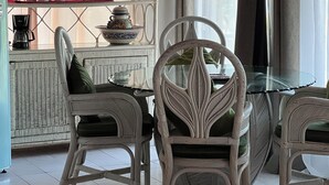 A dining area with wicker chairs and a glass table, a coffee machine, and a decorative shelf with cups and a teapot.