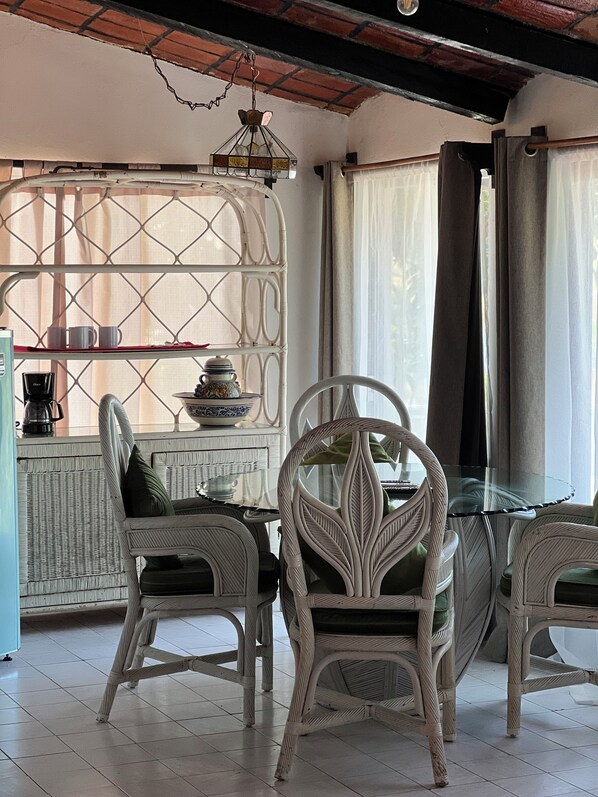 A dining area with wicker chairs and a glass table, a coffee machine, and a decorative shelf with cups and a teapot. - Maison Theurel (Rincón de Guayabitos)