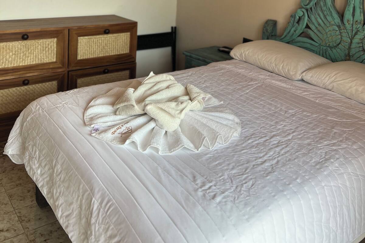 A neatly made bed with a white comforter and a decorative towel arrangement. There is a wooden dresser with woven baskets and a headboard with a leaf design.
