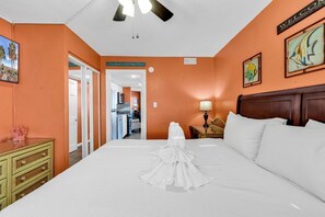 Ein Hotelzimmer mit oranger Wand, einem Bett mit weißen Leinentüchern, einem Holz-Kopfteil, einem Deckenventilator und einem Schild mit der Aufschrift 'Welcome at the Beach'.