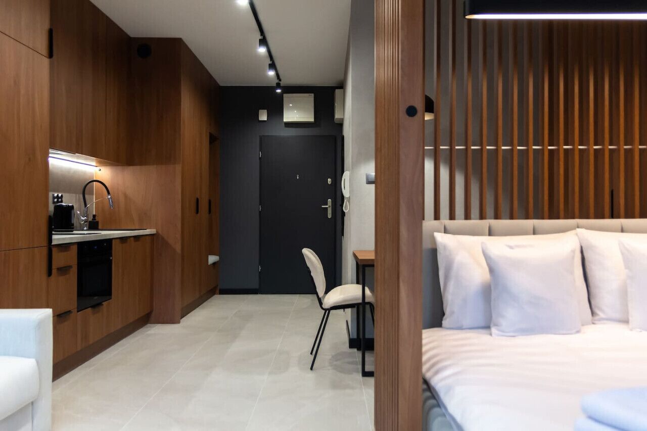 A modern hotel room with a kitchenette, a bed with white bedding, and a wooden panel wall.