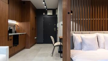 A modern hotel room with a kitchenette, a bed with white bedding, and a wooden panel wall.