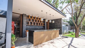 Un espace bar extérieur moderne, doté d’un comptoir en bois, d’étagères en marbre et de luminaires suspendus.