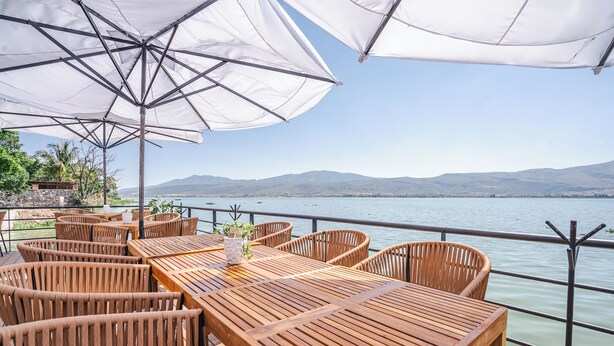 Une terrasse en bois, agrémentée de tables et de chaises, qui offre une vue sur une étendue d’eau paisible et sur des collines lointaines.