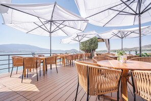 A deck with wooden tables and chairs, white umbrellas, and a view of the sea.