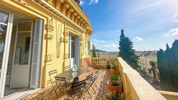 Um edifício amarelo com detalhes arquitetônicos elaborados, um pátio com azulejos, mesa e cadeiras, e vista para a cidade ao fundo.
