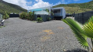 A modern house with a gravel driveway, surrounded by tropical plants and a mountain backdrop.