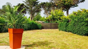 Ein Garten mit einem großen, orangefarbenen Pflanzgefäß, einer saftigen Rasenfläche und verschiedenen Bäumen und Sträuchern.