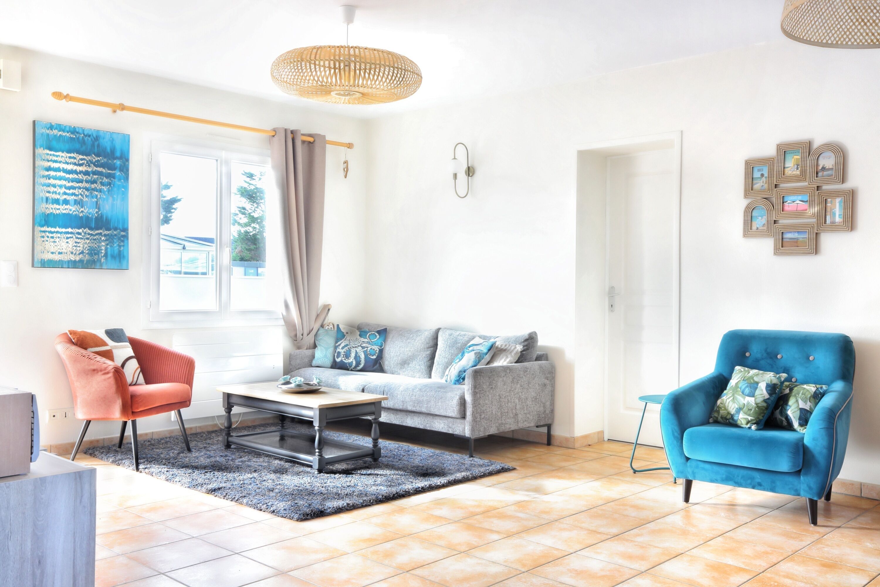 A living room with a grey sofa, a blue armchair, a red chair, and a coffee table. There is a large window with curtains, a wall-mounted light fixture, and a framed picture collection on the wall.