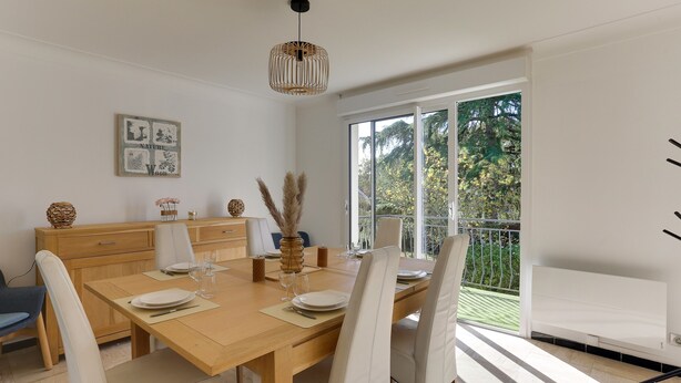 A dining area with a wooden table set for four, white chairs, a vase with pampas grass, and a window with a view of greenery. - Arthur - Appt pour 6 avec jardin partagé (Bruz)
