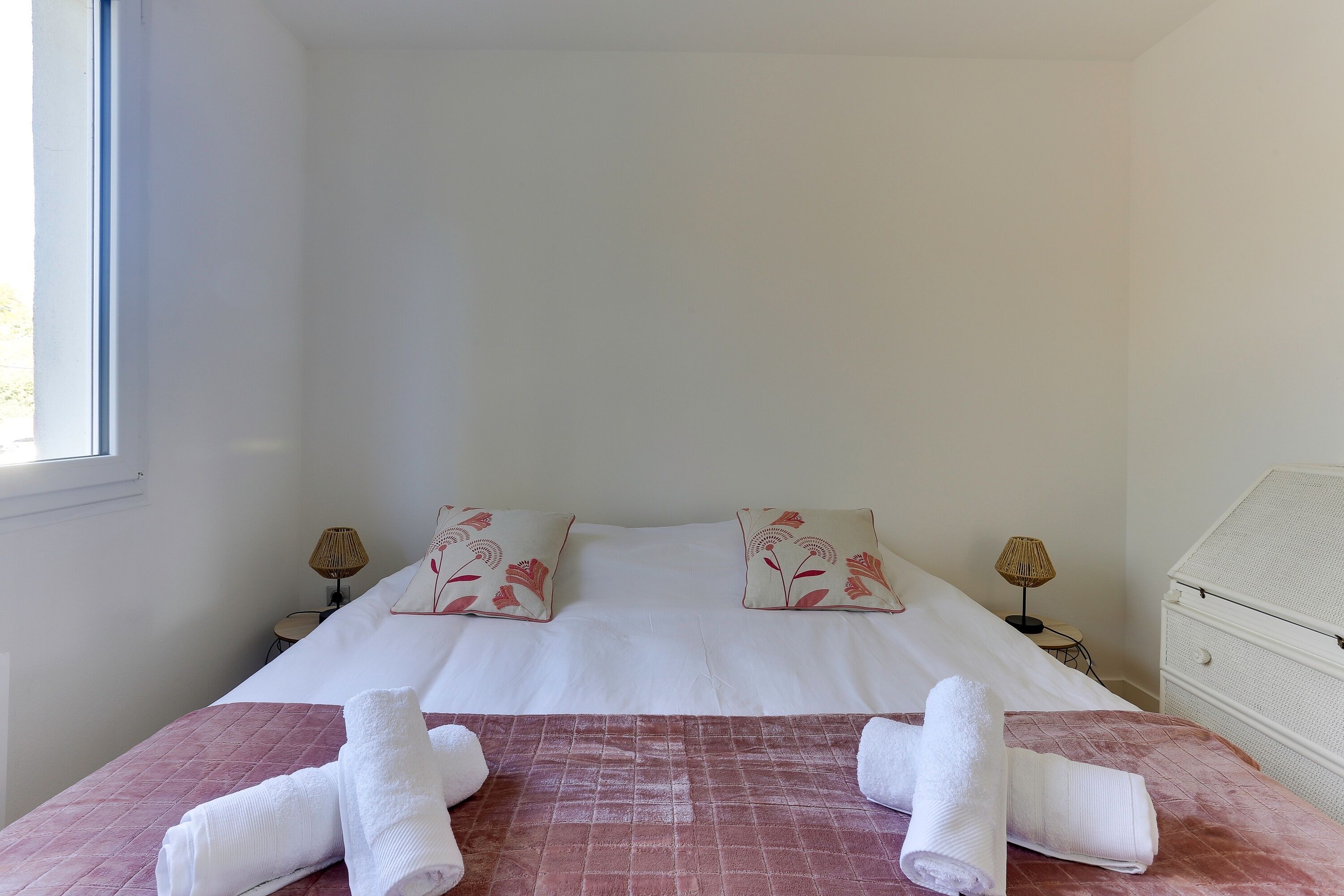 Un lit bien fait, avec des draps blancs et des coussins à motifs floraux, une couvre-lit rose matelassée et deux serviettes enroulées posées sur le lit.