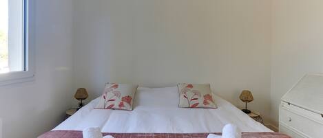 Un lit bien fait, avec des draps blancs et des coussins à motifs floraux, une couvre-lit rose matelassée et deux serviettes enroulées posées sur le lit.