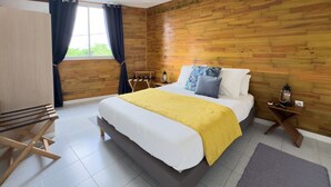A bedroom with a wooden wall, a bed with white and yellow bedding, a wooden nightstand with a lantern, and a chair. - Ti Arums - Appt climatisé vue mer (Les Trois-Îlets)