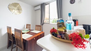 A dining area with a wooden table and chairs, a decorative leaf on the wall, and a window with curtains. - Ti Arums - Appt climatisé vue mer (Les Trois-Îlets)