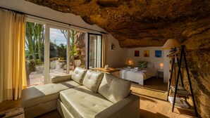 Una sala de estar moderna con un amplio sofá beige modular, una mesa de comedor y una cama en un espacio tipo cueva con paredes de roca natural.