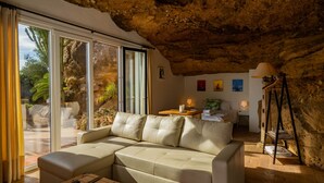 Una sala de estar moderna con techo de piedra, un sofá color beige, una mesa de centro de madera y un amplio ventanal corredero que da a un área exterior con plantas.