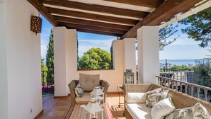 Un patio cubierto con mobiliario de mimbre, un tapete estampado y vistas al mar y la vegetación.