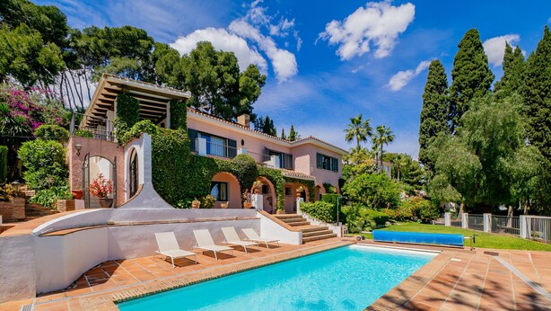 Una piscina con bordeado de baldosas, zona cubierta y un edificio con patio y escalera.