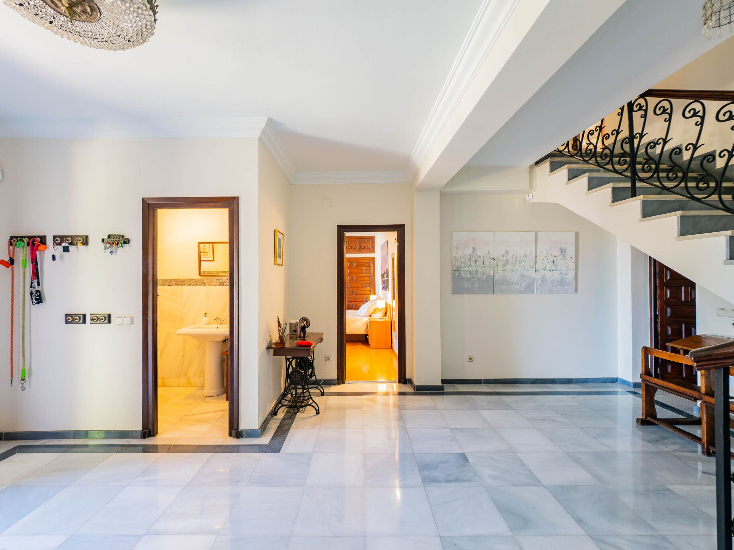 A spacious hallway with a marble floor, a staircase with a wrought iron railing, and a view into a bedroom with a bed and a nightstand.