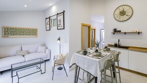Una sala de estar moderna con un sofá, una mesa de comedor para cuatro personas y una zona de cocina con un reloj y estanterías.