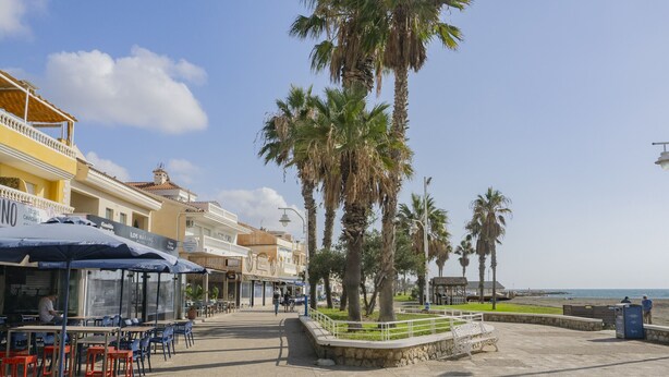 A coastal promenade with outdoor seating, palm trees, and a beach in the background.