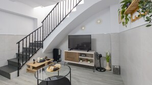 A modern living room with a staircase, a flat-screen TV, a glass coffee table, and a potted plant.