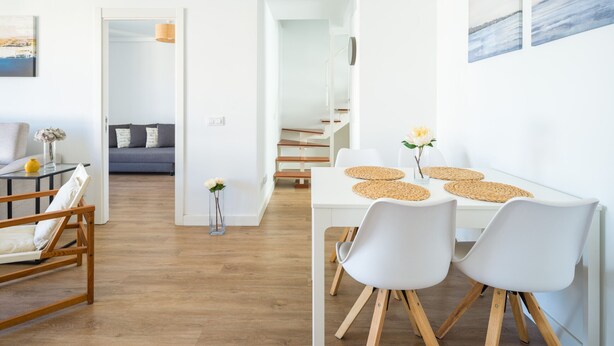 Un interior moderno con un comedor que incluye una mesa blanca, dos sillas blancas y un banco de madera. Una escalera conduce a un nivel superior.