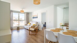 Un moderno salón con sofá, mesa de comedor y sillas. Hay un ventanal con cortinas, una lámpara colgante y un cuadro en la pared.