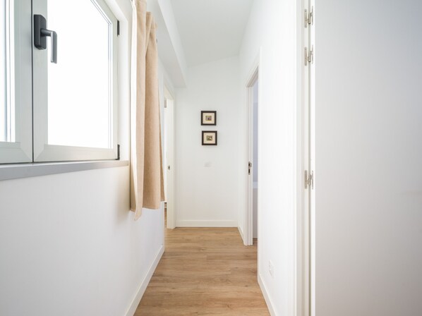 A narrow hallway with white walls, a wooden floor, and a window on the left.