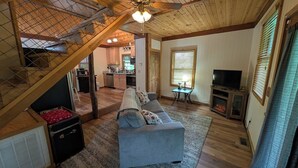 A spacious living area with a sofa, a television, and a fireplace. A kitchen is visible in the background. - Quaint Cabin w/ Elevated Back Deck Among the Trees (Laurelville)