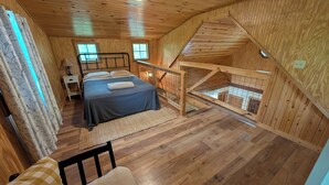 A cozy wooden cabin bedroom with a bed, a nightstand, a window, and a loft area. - Quaint Cabin w/ Elevated Back Deck Among the Trees (Laurelville)