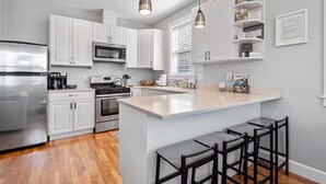 Una cocina moderna con armarios blancos, electrodomésticos de acero inoxidable, una isla central con taburetes y lámparas colgantes.