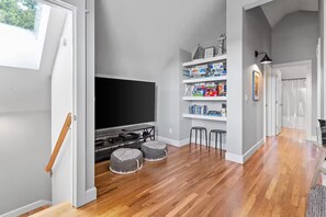 A room with a flat-screen TV, wooden flooring, a bookshelf with various items, and a window with a view of trees.