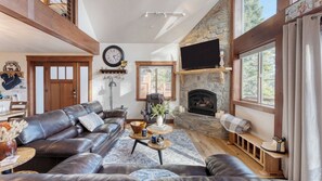 Un salon spacieux avec un canapé en cuir, une cheminée en pierre, un téléviseur fixé au mur et une grande fenêtre donnant sur les arbres.