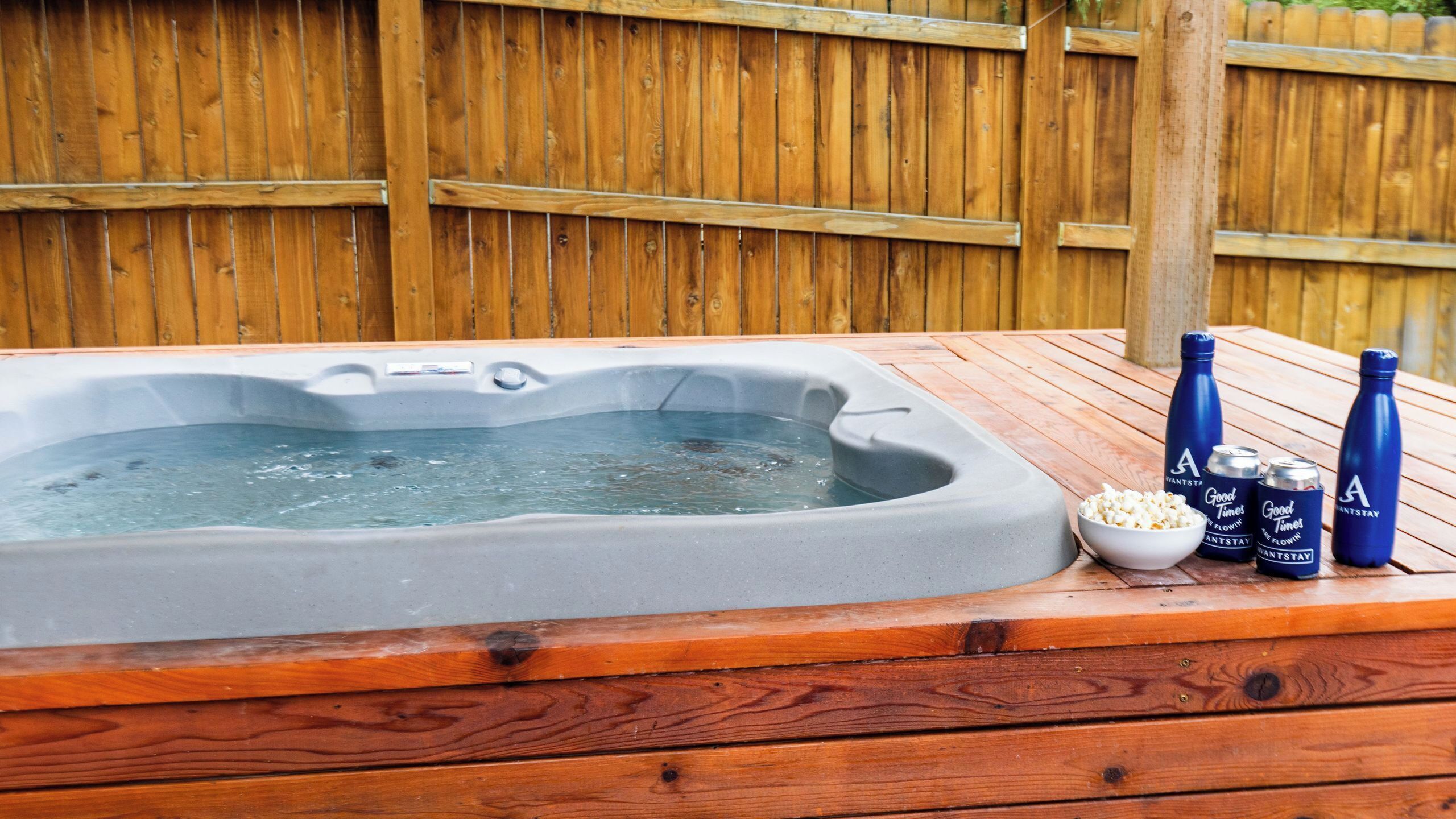 A hot tub with a wooden deck, a bowl of popcorn, and three bottles labeled 'Good Times' on a wooden fence background.