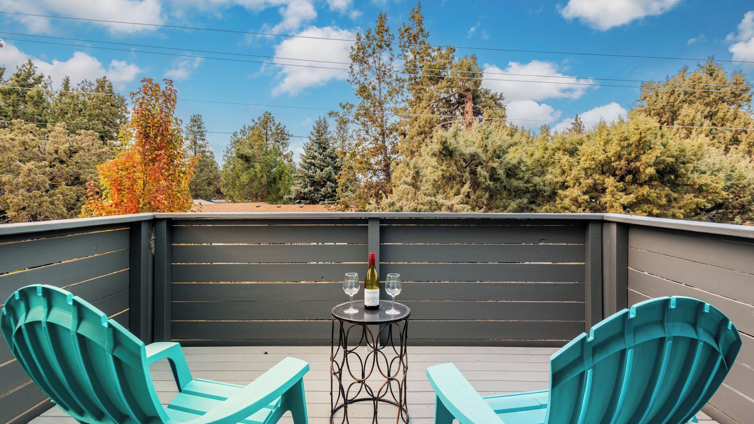 A covered outdoor seating area with two turquoise chairs, a small round table with a bottle and glasses, and a view of trees and sky.