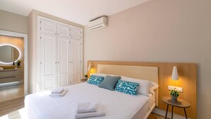 A neatly made bed with white linens and two pillows, a bedside table with a lamp, and a wall-mounted air conditioner.