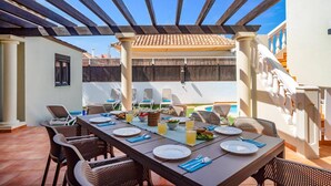 An outdoor dining area with a pergola, dining table, chairs, and a pool in the background.
