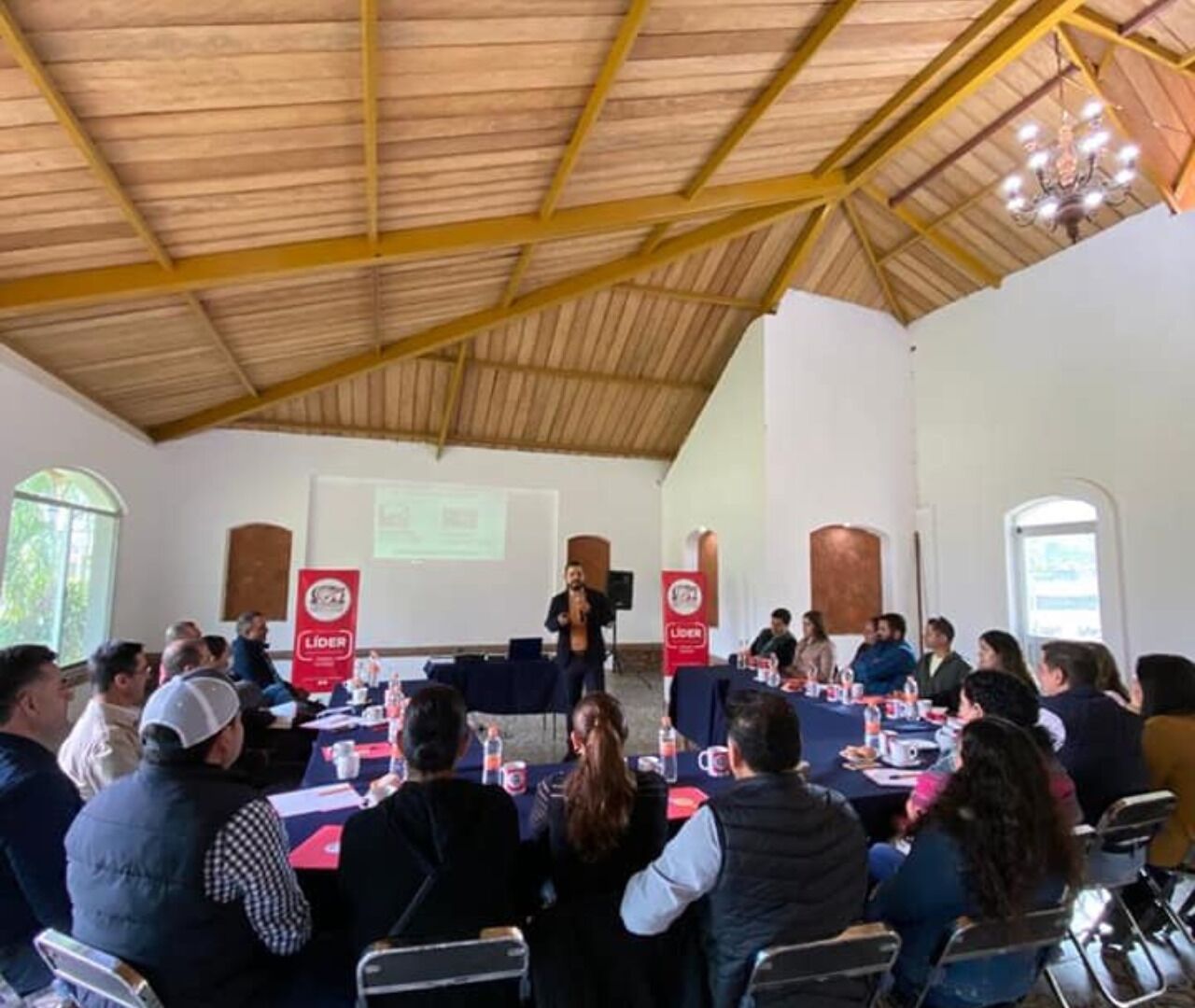 Un salón de conferencias con techo de madera, un orador al frente y asistentes sentados en mesas con placas de identificación.