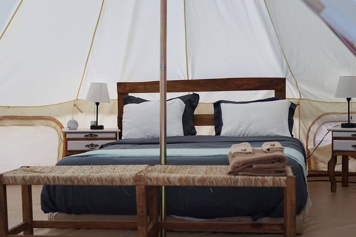 A tented accommodation with a bed, bedside tables, lamps, and a bench with towels.
